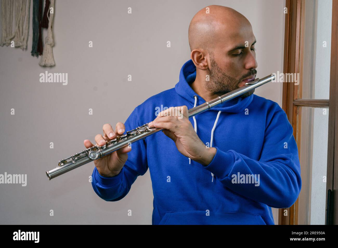 portrait of young latino man in blue, with beard and bald head standing at home playing flute with inspiration Stock Photo