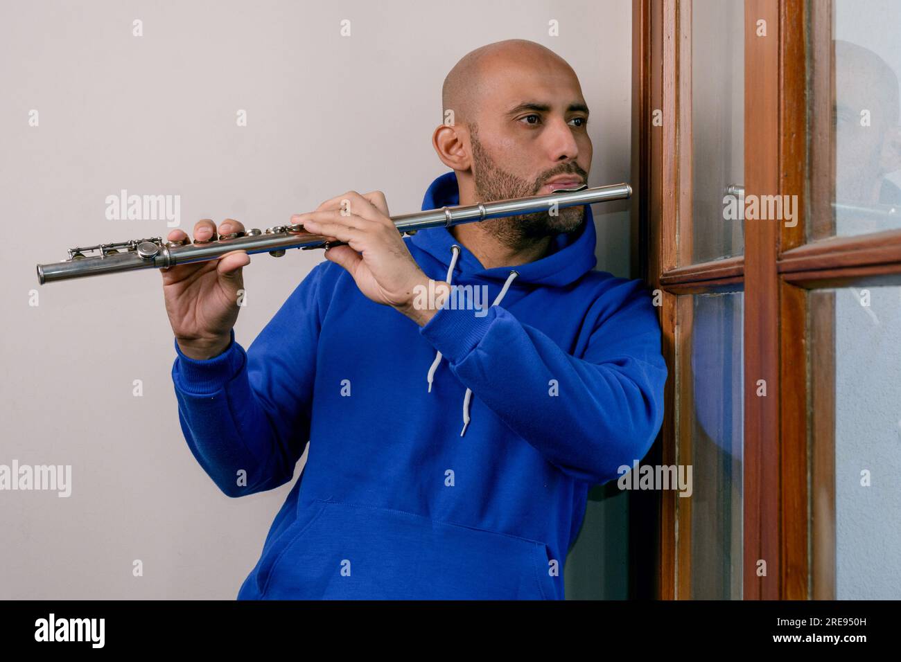 portrait of young latino man at home alone playing the flute, looking out the window inspired and melancholic. Stock Photo