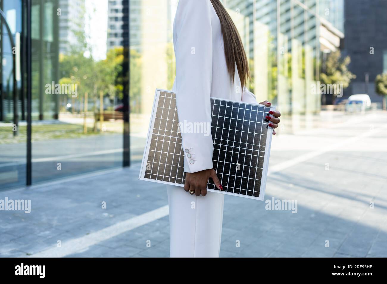 Side view of crop anonymous businesswoman in white blazer standing holding on street with portable solar panel on sunny day Stock Photo