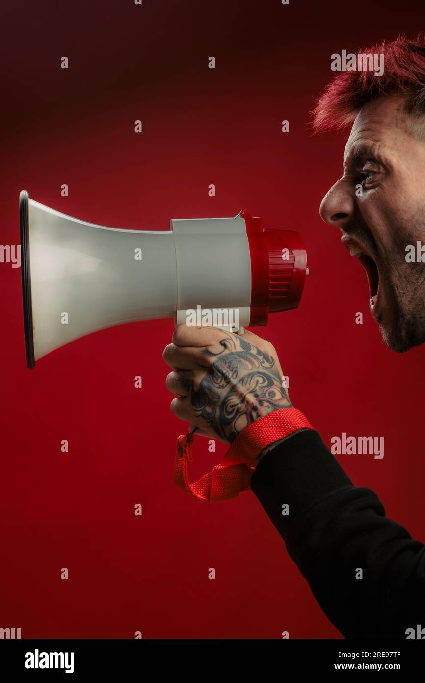 Side view of crop anxious male with tattoo hand and red hair shouting over megaphone on red studio background Stock Photo