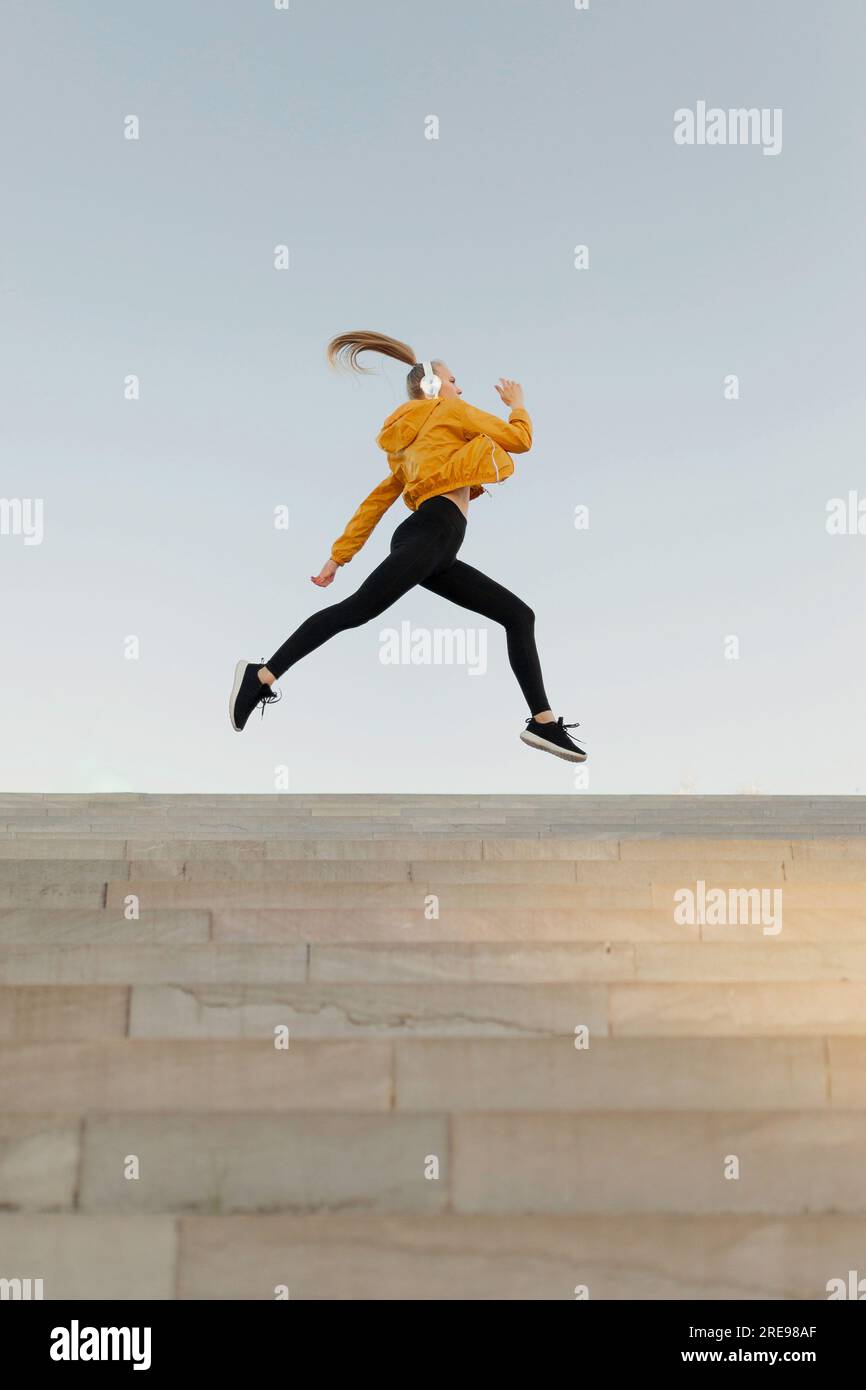 Young athletic caucasian woman wearing headphones and sport outfit, jumping on stairs outdoors Stock Photo