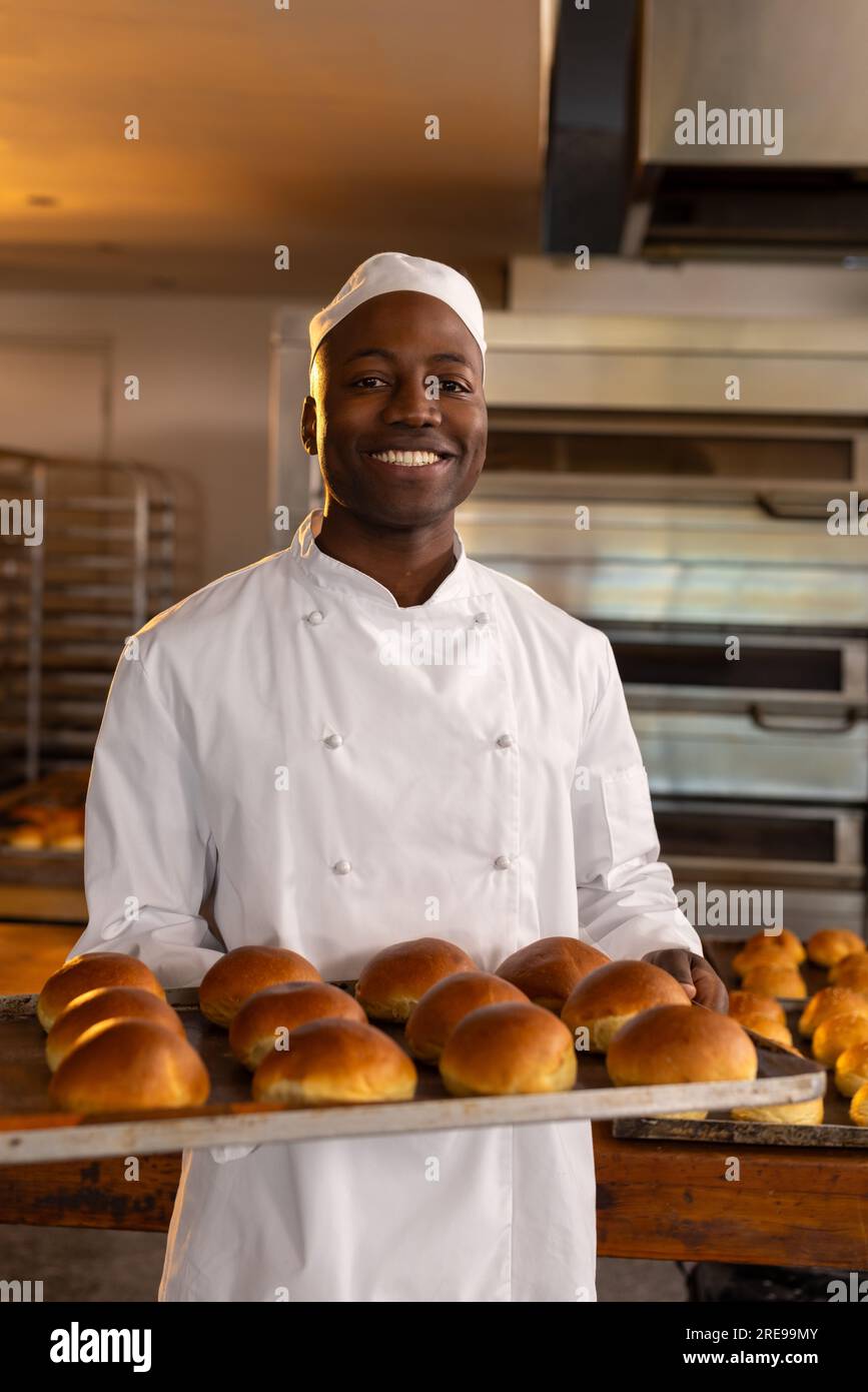 Portrait of happy african american male baker in bakery kitchen presenting fresh rolls Stock Photo