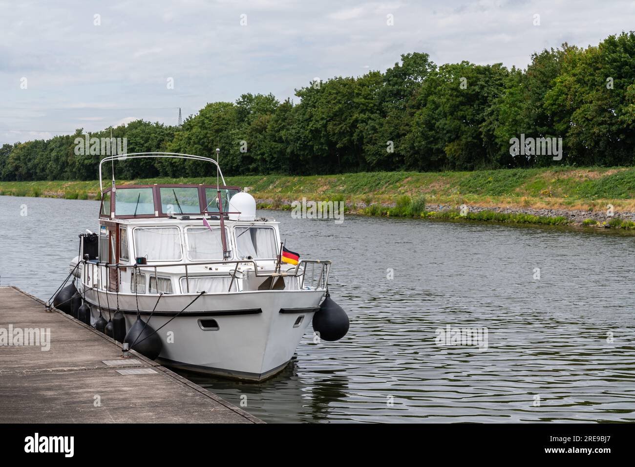 A small boat with a German flag on the bow is moored to a pier on the river. Stock Photo