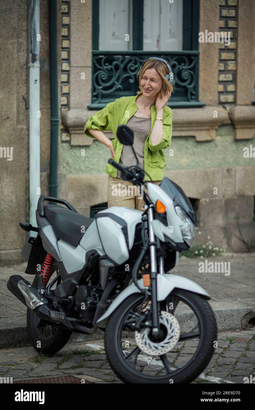 A woman wearing headphones standing in the street near a motorcycle. Stock Photo