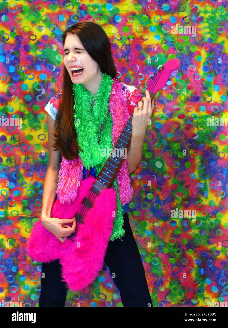 Laughing hillariously, female teen pretends to strum on the stuffed, fuzzy, base guitar.  A tye-dye background adds vibrant color and fun. Stock Photo