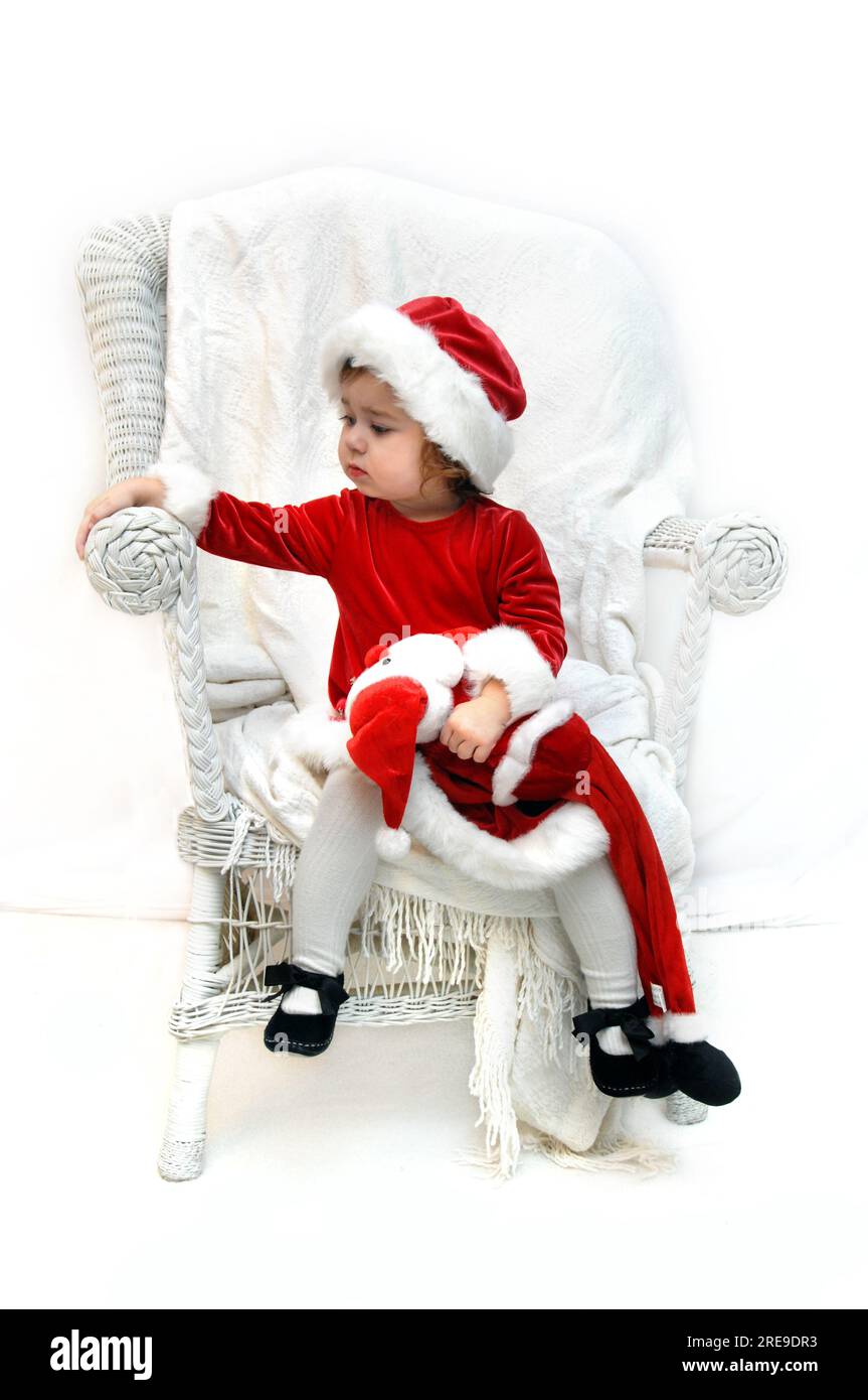 Little girl, dressed in a santa claus costume, sits and waits on Santa.  She is sitting ina white chair in a white room. Stock Photo