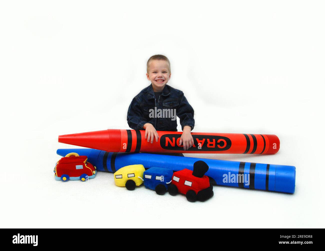 Little boy is sitting in a white room surrounded by large crayons and toys.  He is brimming with joy. Stock Photo