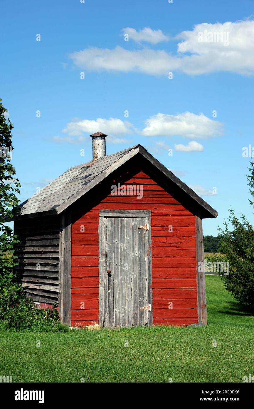 Old, red, wooden smokehouse has cracked and peeling paint. Stove pipe