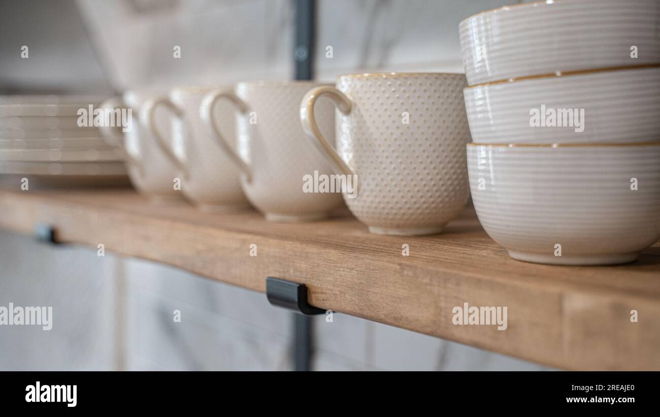 Modern wooden Kitchen shelf with white textured cups, bowls and plates. Light tile backsplash in the background Stock Photo