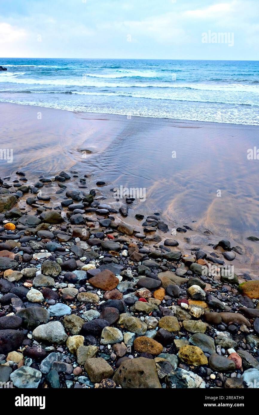 Stony beach after sunset, wet sand at the ebb, surf and remote blue horizon. Soft toned calming seascape. Stock Photo