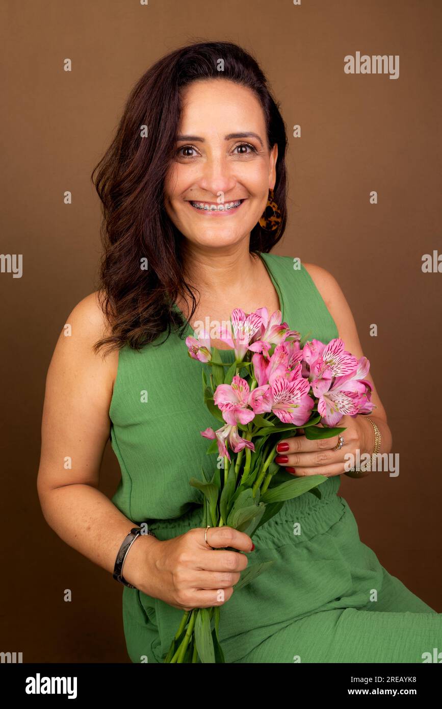 Beautiful woman, wearing green clothes, very happy, holding a bouquet of flowers. Confidence, motivation and maturity concept. Isolated on brown backg Stock Photo