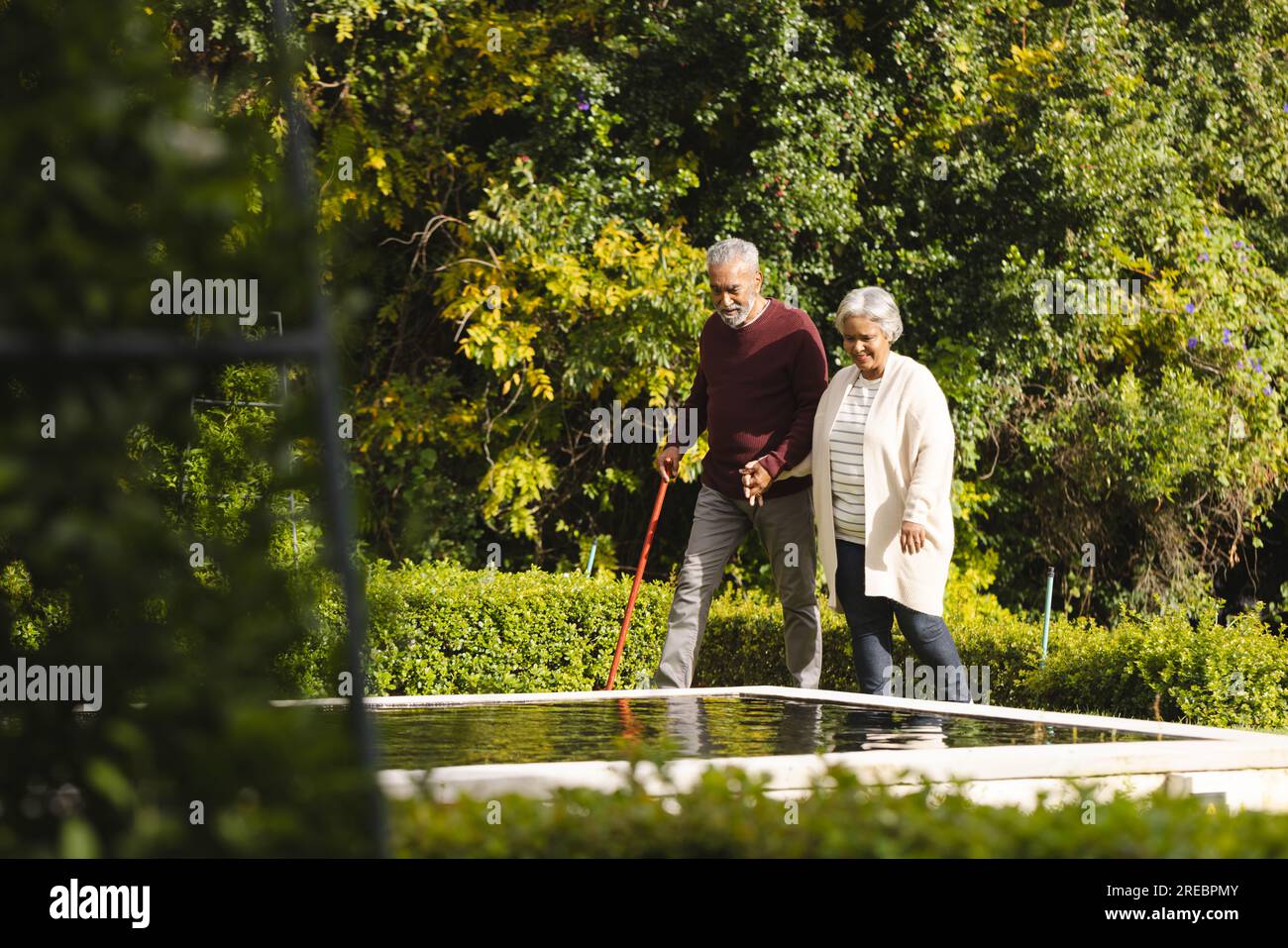 Happy senior biracial couple holding hands and walking with cane in garden at home Stock Photo