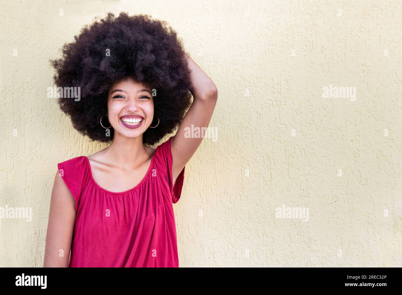 Portrait of positive African American female with Afro hairstyle smiling with raised hand and looking at camera while standing against beige backgroun Stock Photo