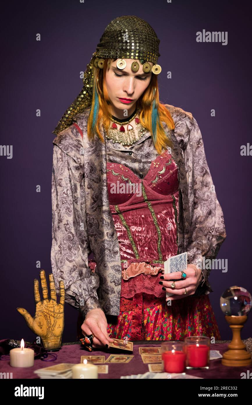 Serious young female fortune teller with long ginger hair in boho style clothes laying out tarot cards standing at table with burning candles and crys Stock Photo