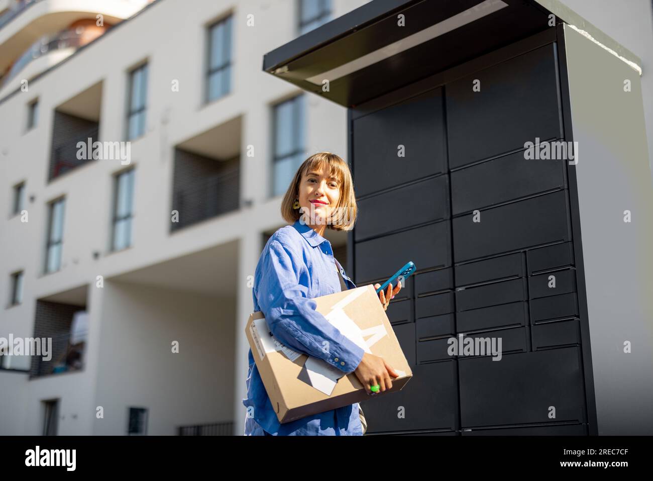 Woman with phone and parcel near automatic post office machine in residential district Stock Photo