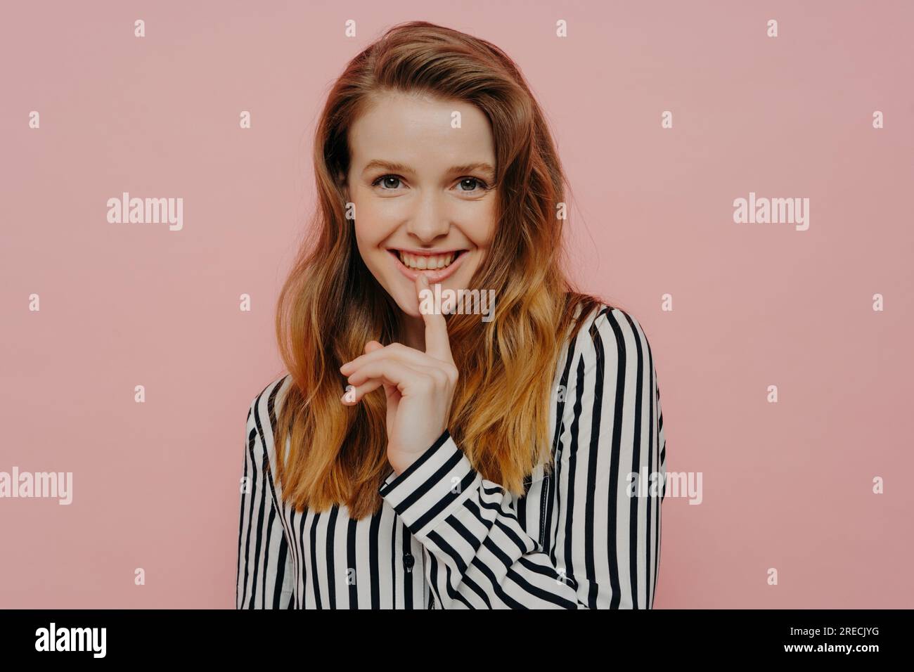 Beautiful, happy young woman in stripy black and white blouse, touching lips with pointer finger, posing in studio on light pink background. Stock Photo