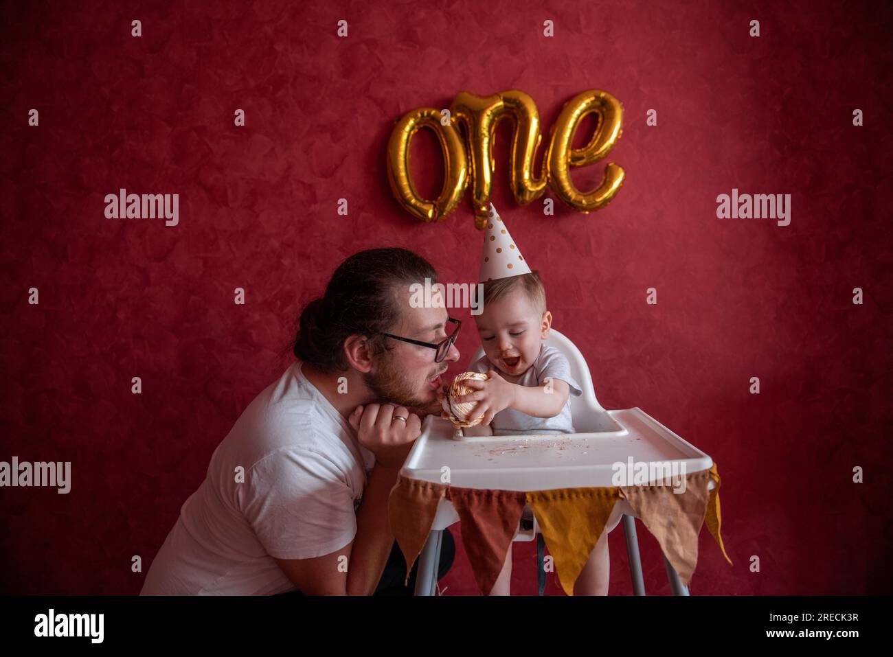 Father celebrates with little son his first birthday. Man fooling around with boy, eats holiday cake against red wall background with One foil balloon Stock Photo