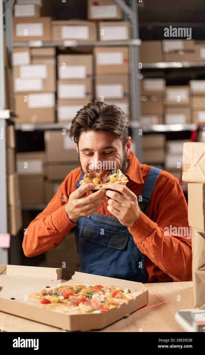 Young warehouse worker eating pizza at lunch at workplace, food at work concept. Closeup Stock