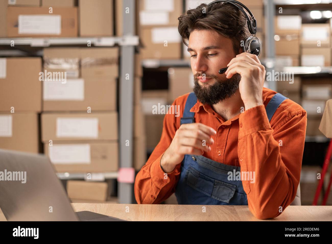 Male worker with headset working in on-site office of a warehouse using laptop computer consulting clients Stock Photo