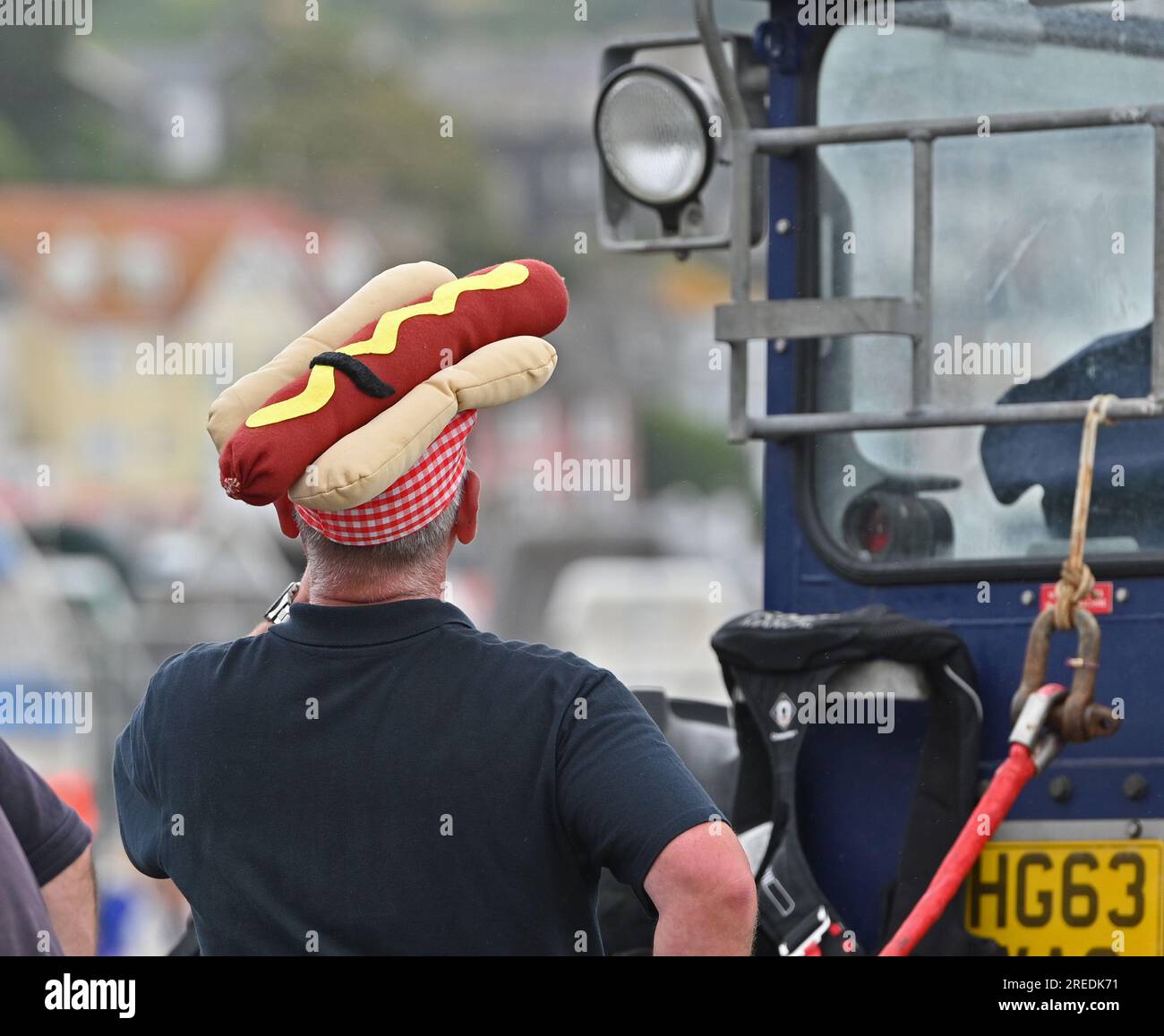 Lyme Regis, UK. 27th July, 2023. On a warm and damp afternoon, Dave a crew member of the RNLI was seen with a Hotdog on his head, calling out Hotdogs for sale raising funds for the RNLI Lifeboat week at Lyme Regis East Devon. Picture Credit: Robert Timoney/Alamy Live News Stock Photo