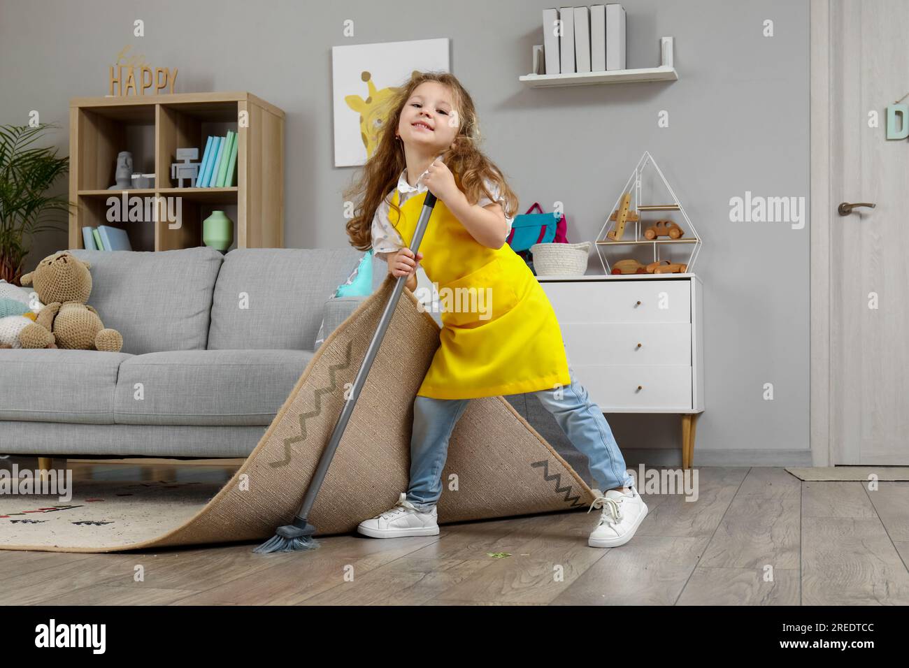 Cute little girl sweeping floor under carpet with broom at home Stock