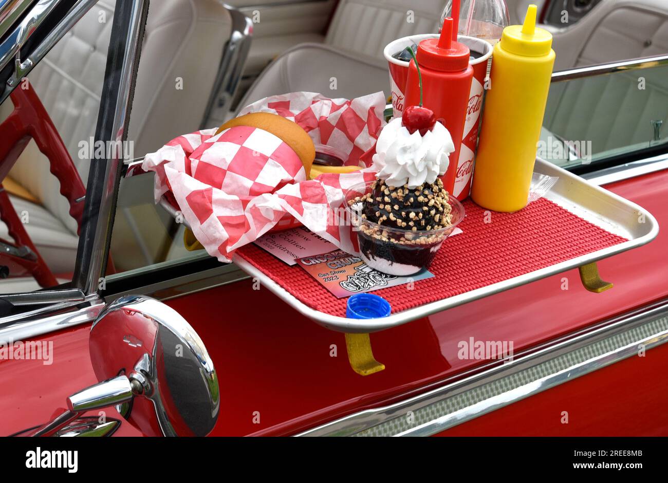 A 1950s Chevrolet convertible on display at a car show features a drive-in restaurant window tray with fast food and drinks. Stock Photo