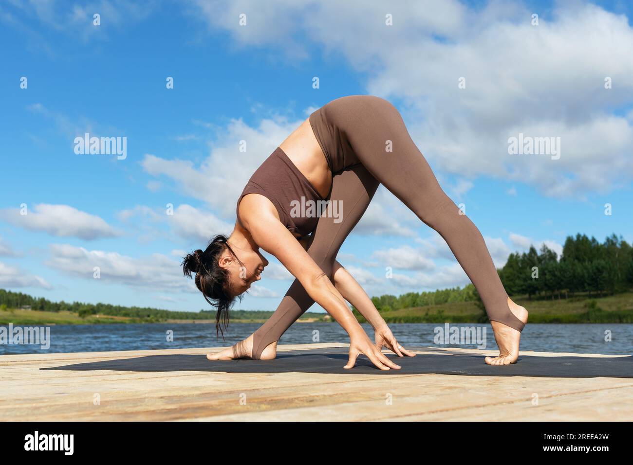 A female yoga practitioner performs a variation of Parshvottanasana, Intense Lateral Stretch Pose, exercising in sportswear on a lakeside on a warm su Stock Photo