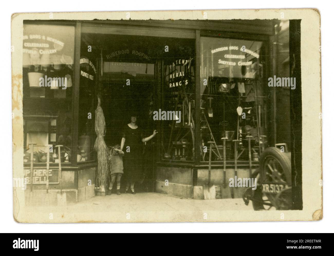 Original 1920's small postcard Maurice A. Clarke Ironmongers / hardware shop - proprietor's wife stands in a doorway with her child. Outside the shop is parked the family's Morris Cowley 'Bullnose' open 2 seater. The rear can just be seen with the letter prefix XR indicating a London registration, U.K. Circa 1925 Stock Photo