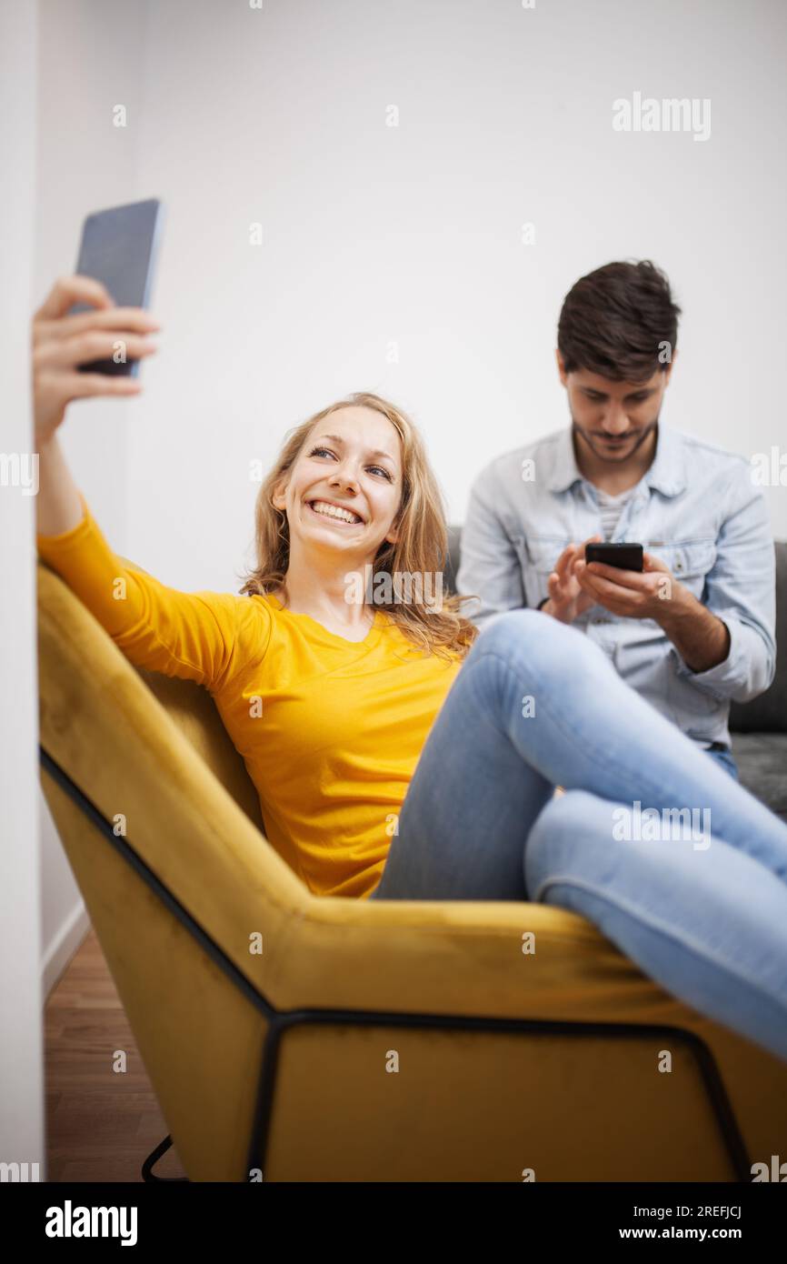 girl at home taking selfie on her smart phone. boyfriend in the backgroung paying no attention. Stock Photo