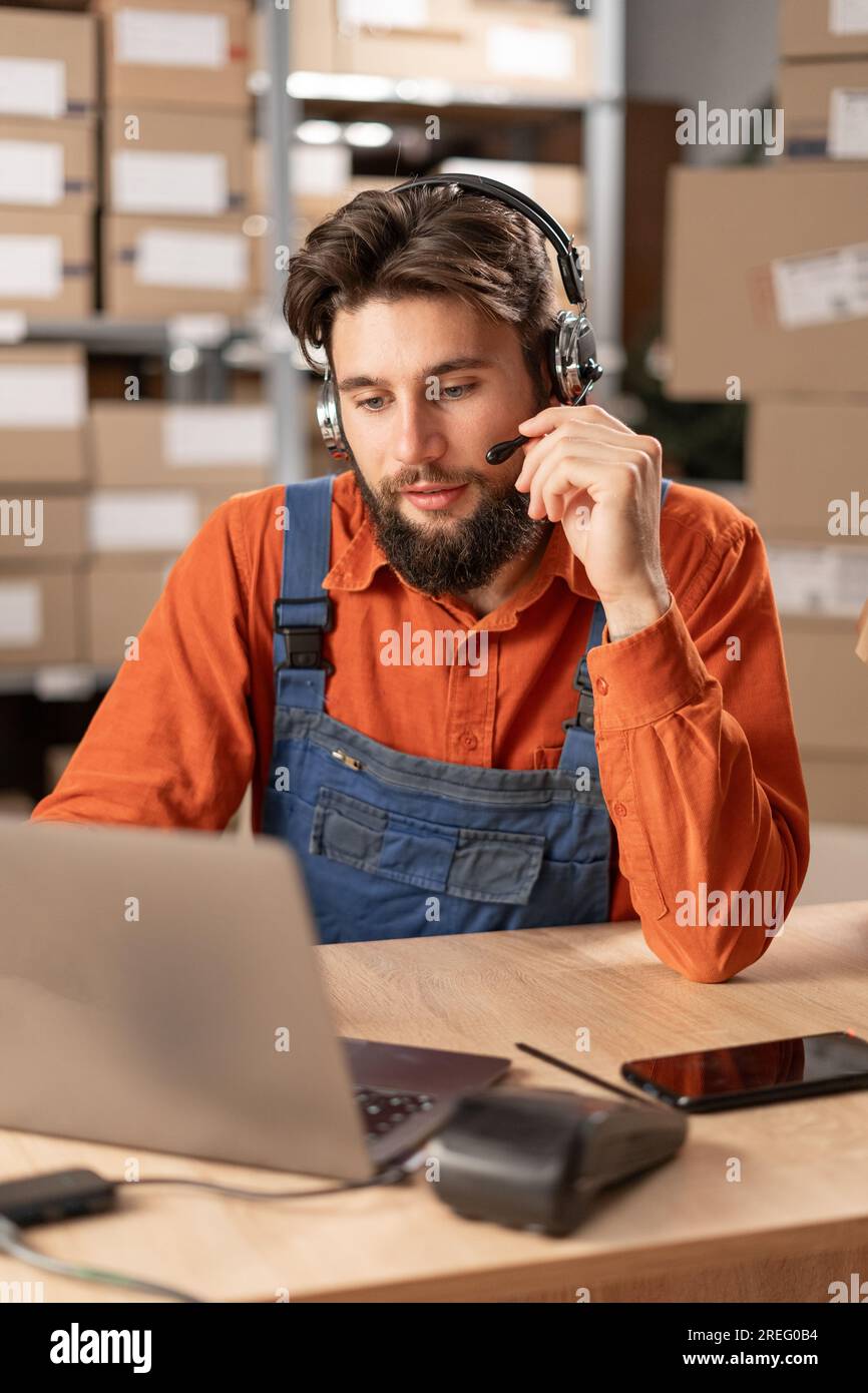 Male worker with headset working in on-site office of a warehouse using laptop computer consulting clients. Stock Photo