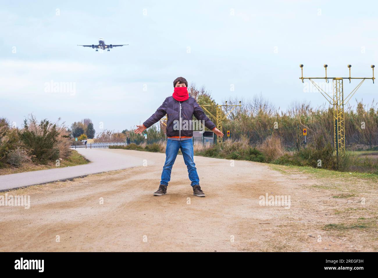 Front view of a boy standing with outstretched arms and wearing a red scarf while an airplane taking off in airport Stock Photo