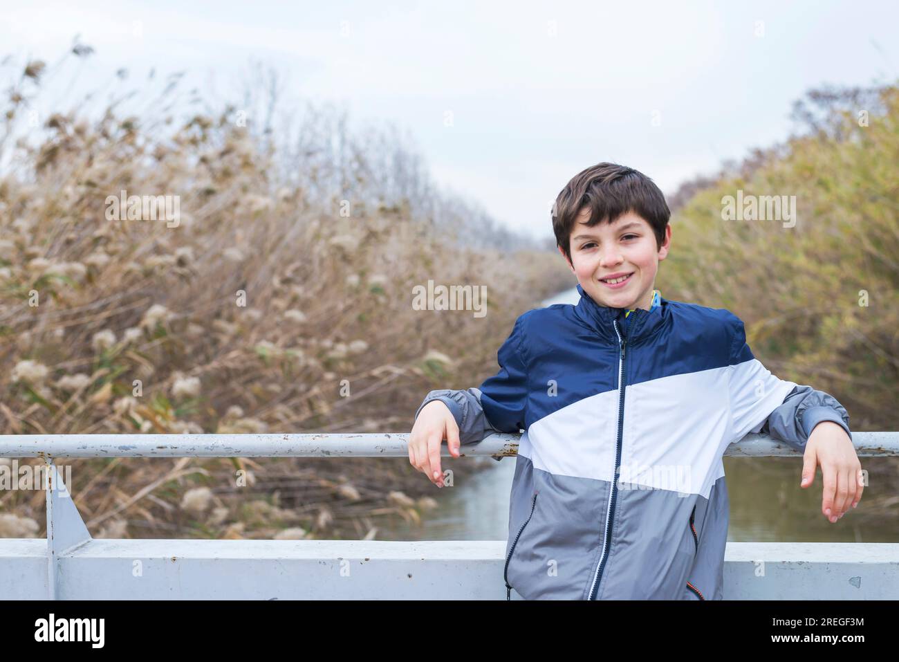 Full length portrait of young teen standing by railing on bridge while looking camera and smiling against river Stock Photo