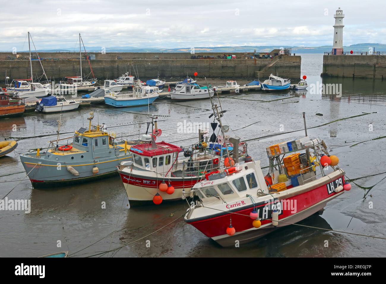 Fishing boats moored at low tide in Newhaven Harbour, Edinburgh, with the harbour lighthouse and views across the Firth of Forth towards Fife Stock Photo