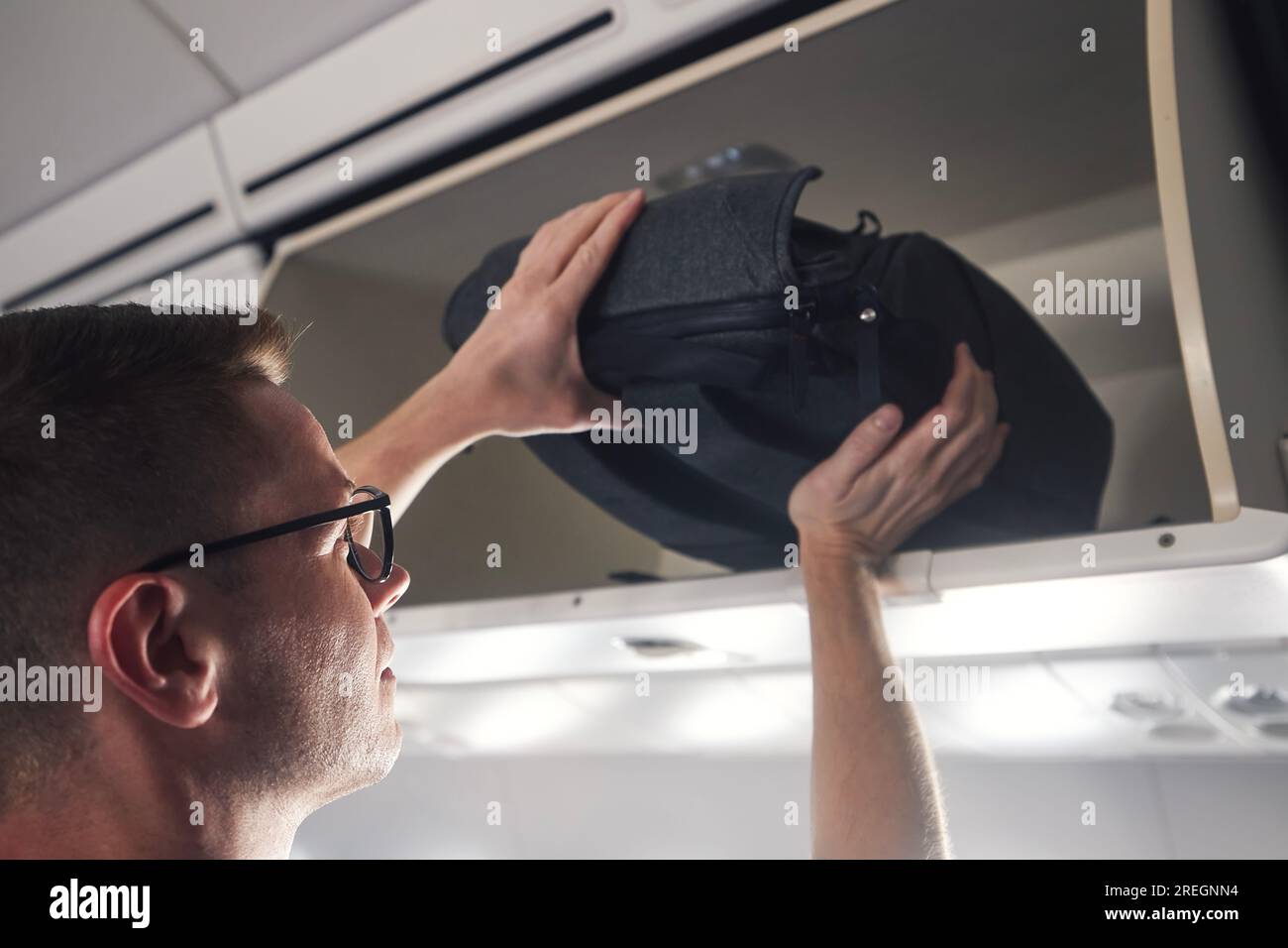 Man with backpack is travel by airplane. Passenger putting hand baggage in lockers above seats of plane. Stock Photo