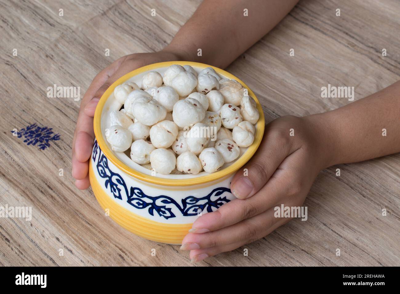 Hand holding beautiful yellow and blue pot containing Makhana or foxnut in brown background. Offering Healthy Food. selective focus on subject Stock Photo