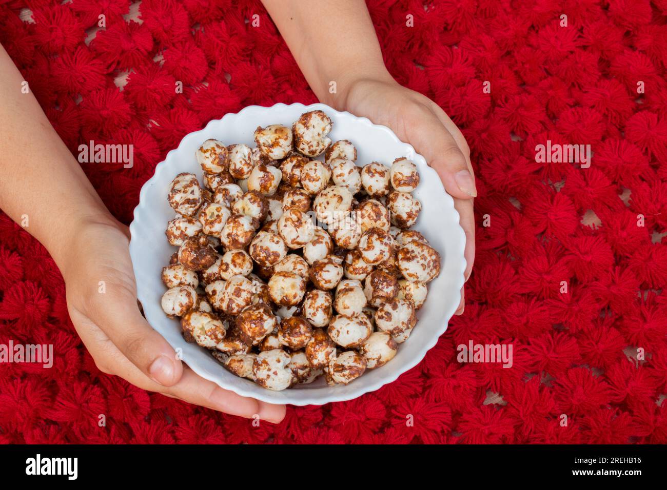 Hand holding white bowl having healthy Chocolate and jaggery coated makhana in beautiful red background. copyspace Stock Photo
