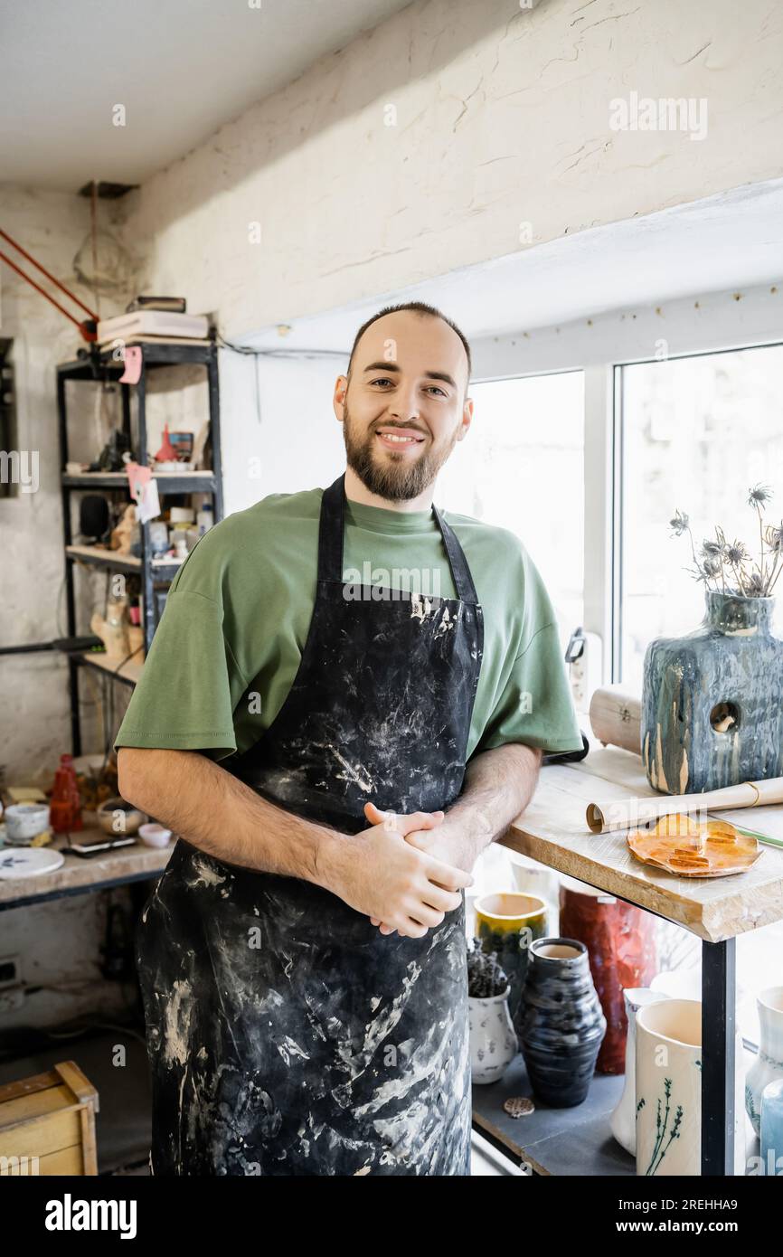 Smiling craftsman in apron looking at camera and standing near vases and window in ceramic workshop Stock Photo