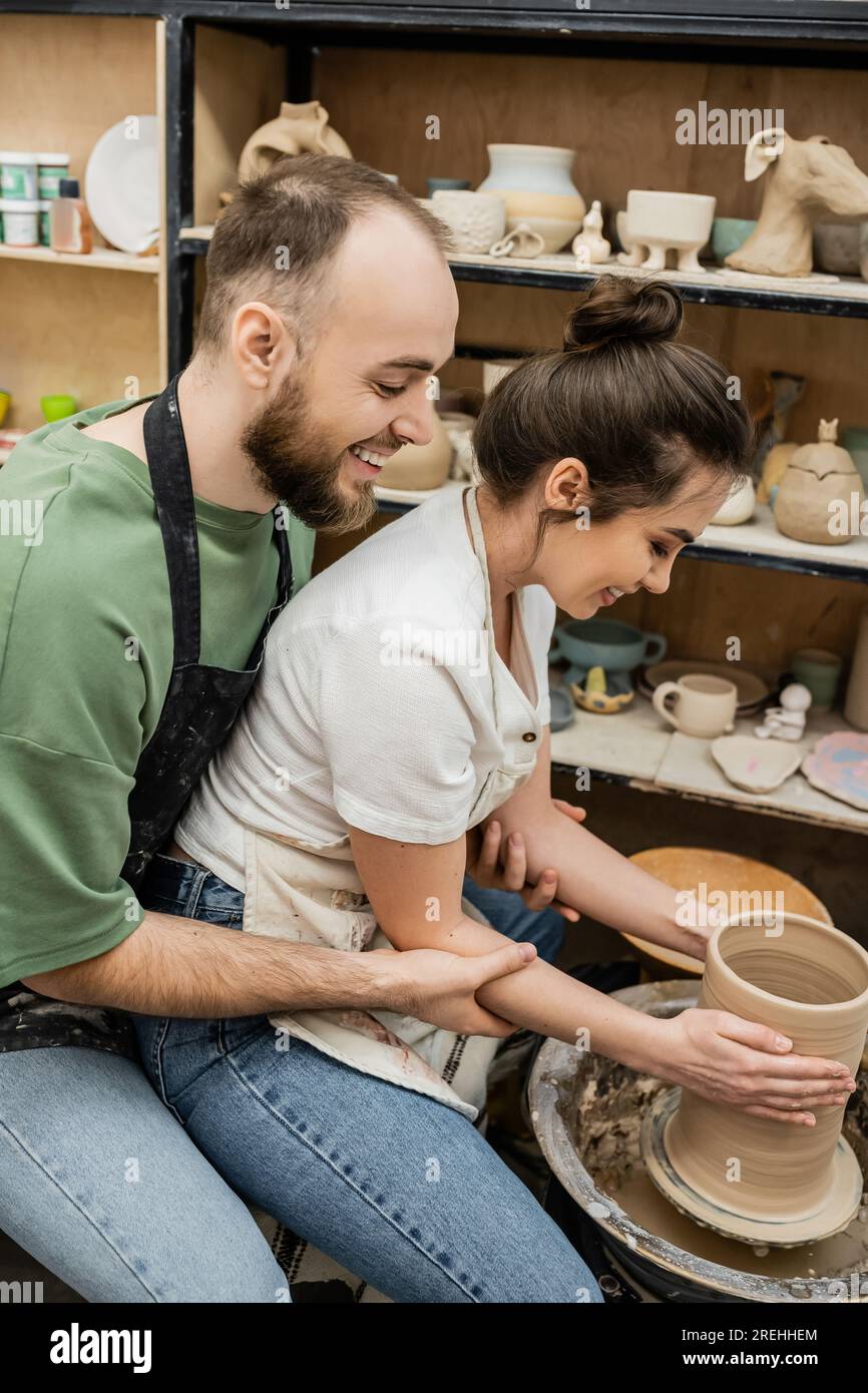 Side view of cheerful romantic artisans in aprons making clay vase on pottery wheel in
