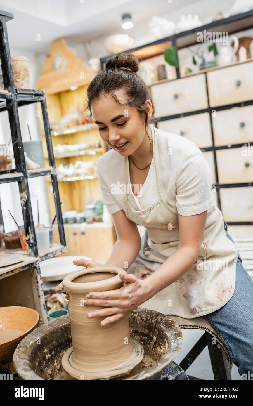 Positive female artisan in apron shaping clay vase on pottery wheel in ceramic studio at