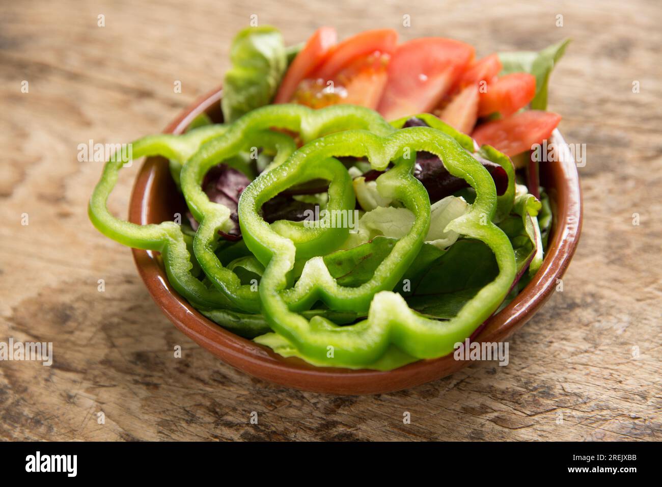 A bowl of mixed salad set on a wooden chopping board. It consists of sliced green bell peppers, tomatoes and baby leaf salad. England UK GB Stock Photo