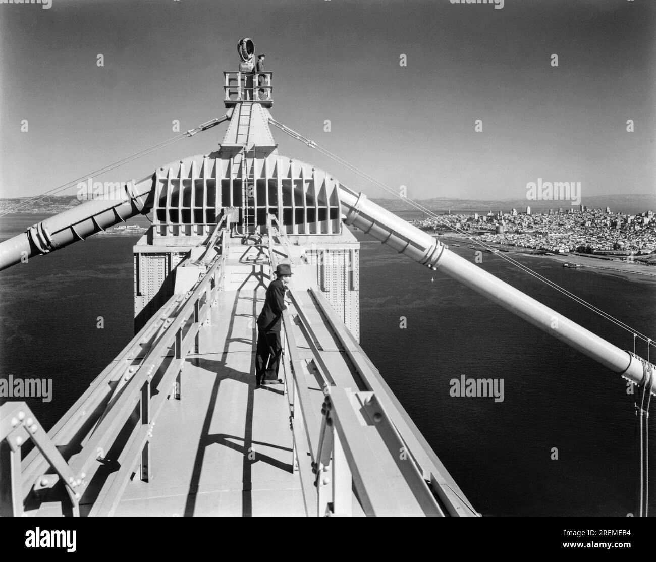 San Francisco, California:   May 29, 1937 A man standing atop one of the towers of the completed and opened Golden Gate Bridge with San Francisco in the background. Another man is at the very top above the saddle by the aviation light. Stock Photo
