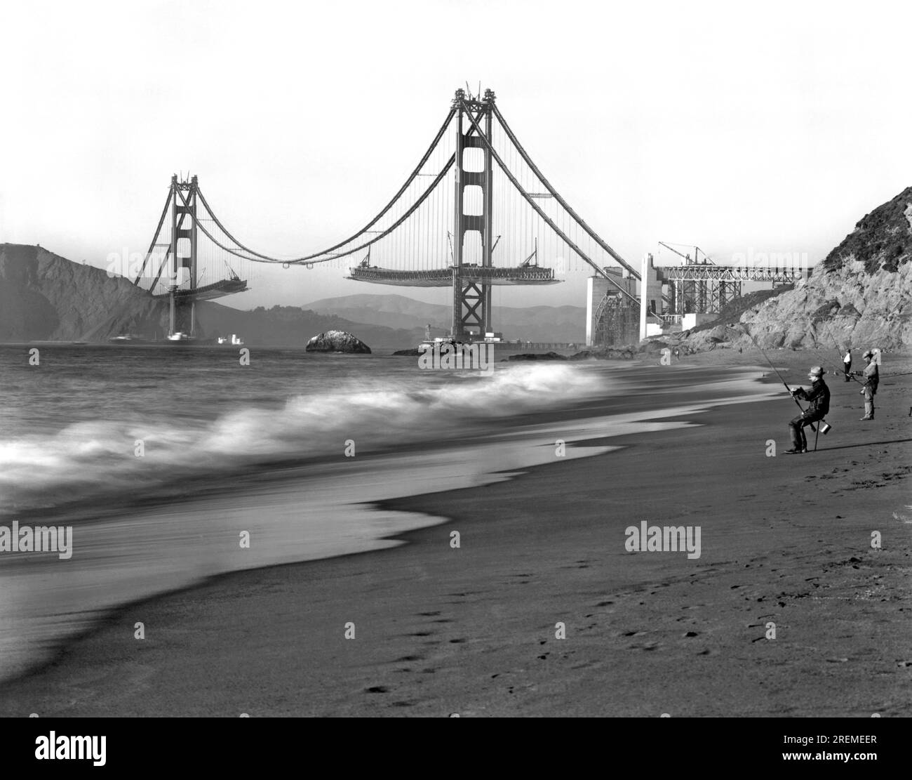 San Francisco, California:   c.1936. Fishermen on Baker Beach enjoy the view of the Golden Gate Bridge under construction. Stock Photo