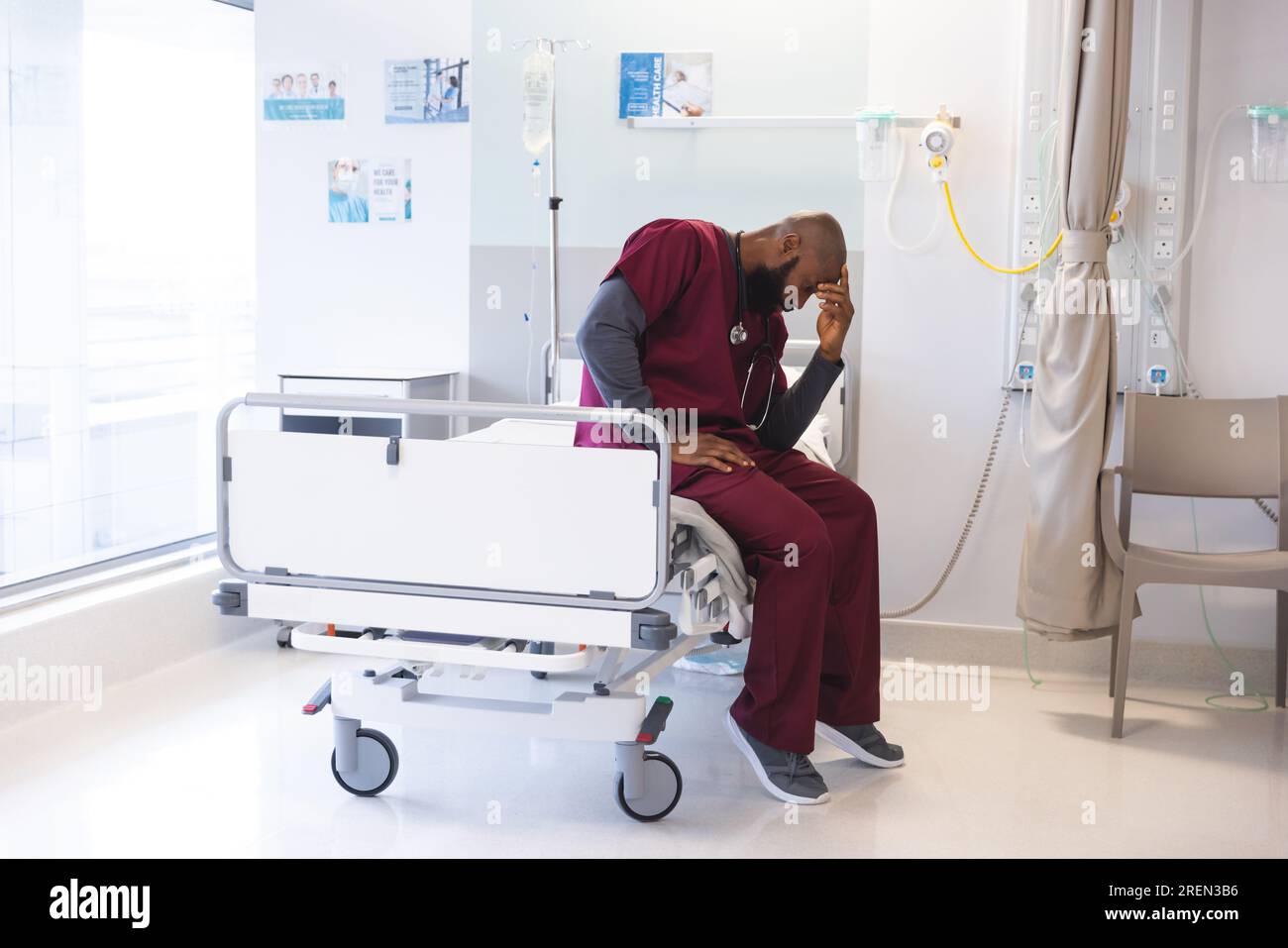 Sad african american male doctor wearing scrubs sitting on bed at
