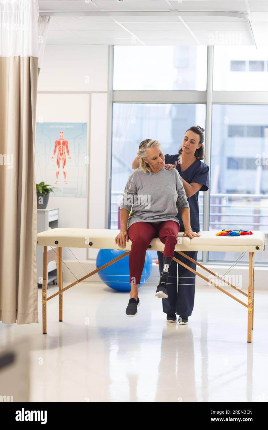 Caucasian female physiotherapist and senior woman with artificial leg massaging back at hospital Stock Photo