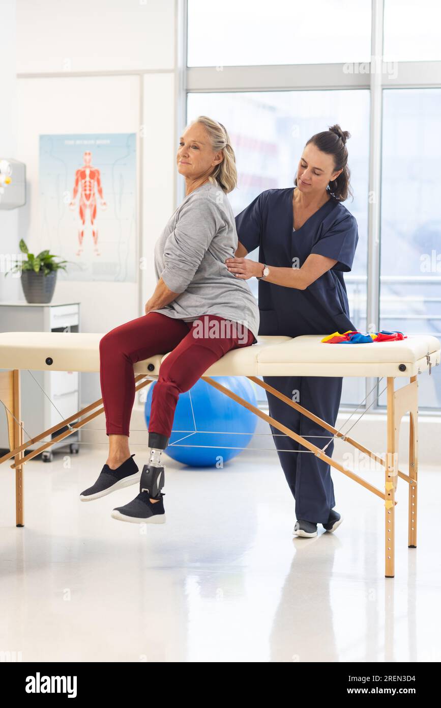 Caucasian female physiotherapist and senior woman with artificial leg massaging back at hospital Stock Photo