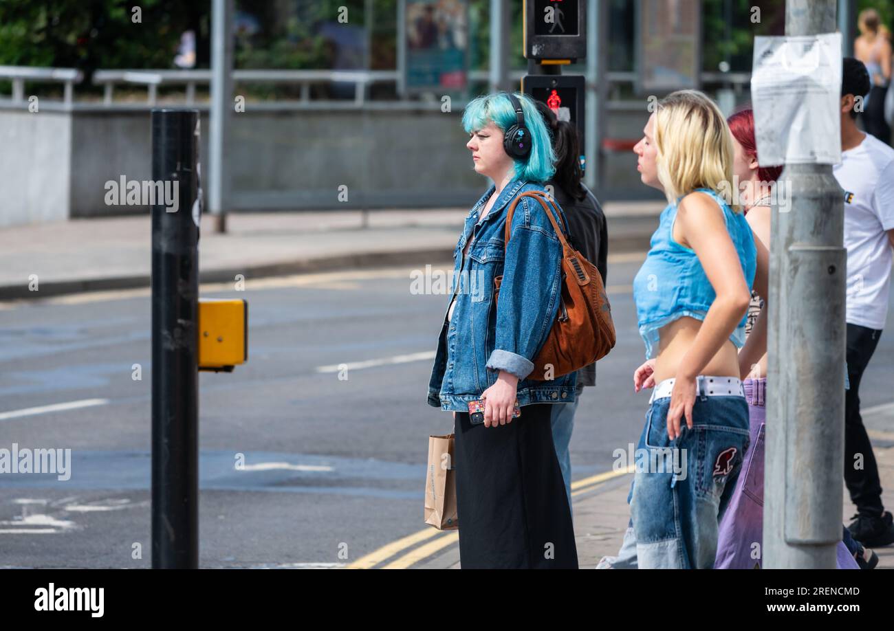 Young woman fashionably dressed, trending, with green hair wearing headphones waiting to cross a road in Brighton & Hove, UK. Stock Photo