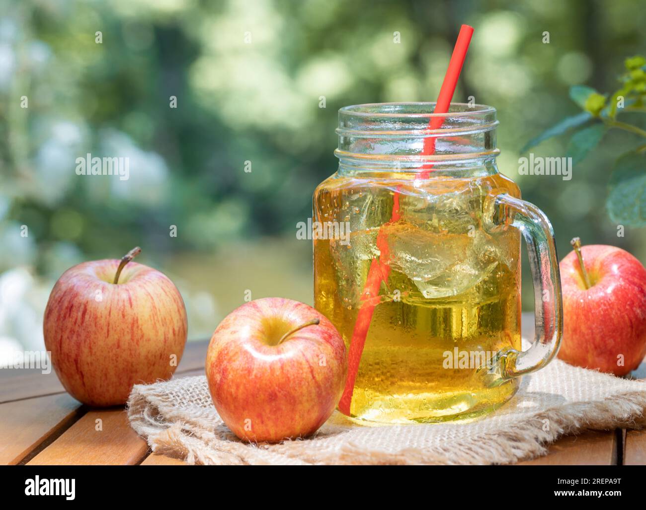 Cold apple juice in glass jar and fresh red apples outdoors on wooden patio table with nature background Stock Photo