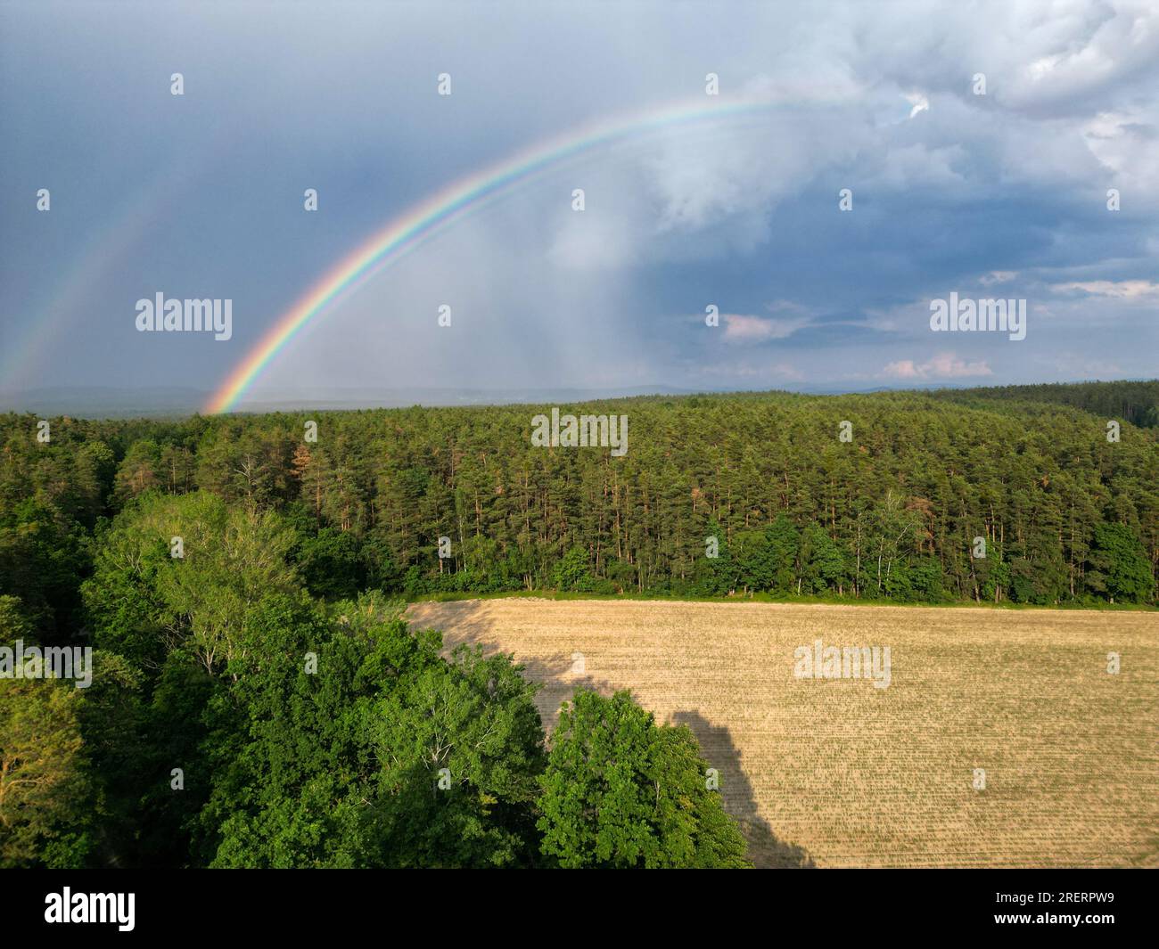 Beautiful double rainbow arching over field and forest with dark clouds and rain in the background Stock Photo