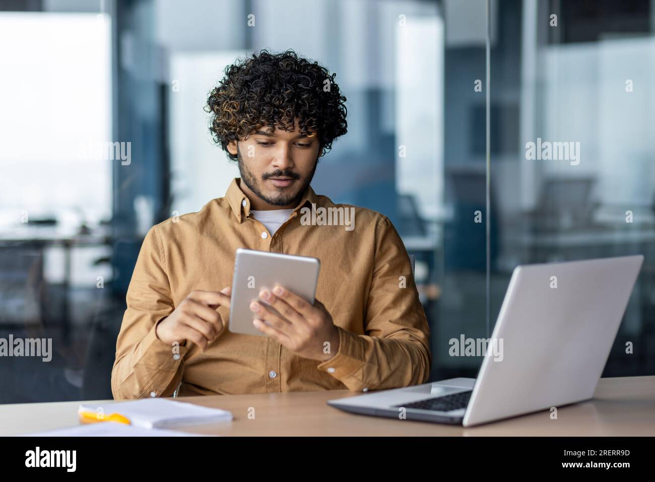 Serious focused businessman working with tablet computer sitting at desk inside office, hispanic thinking with laptop, man solving technical problem, using online application. Stock Photo
