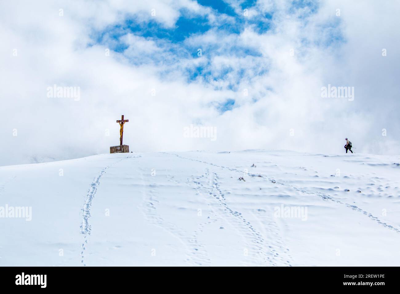 Lonely hiker approaching a statue of Jesus on the cross on top of a snowy mountain with blue sky surrounded by clouds Stock Photo